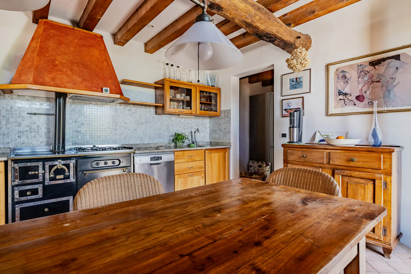 Rustic kitchen with a large wooden table, black stove, orange hood, and exposed wooden beams on the ceiling.