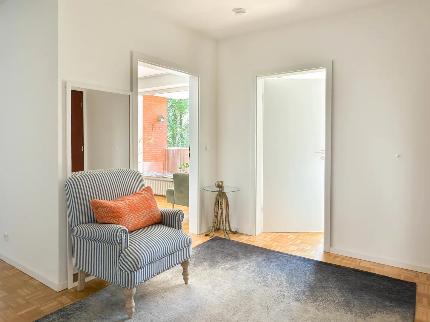 Bright, white-walled room with striped armchair, orange pillow, and blue rug. Open doorways lead to other rooms.