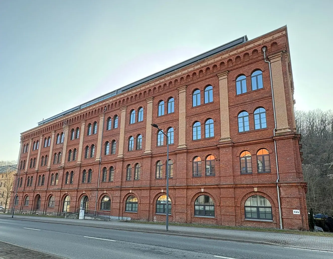 A long, red brick building with arched windows and tan pillars, next to a street.