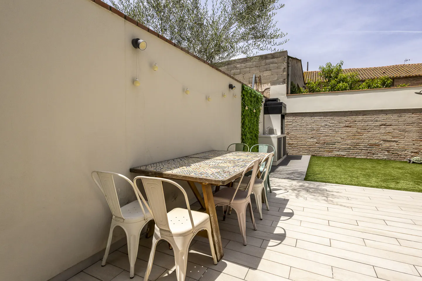 Outdoor patio with a tiled dining table, metal chairs, string lights on a cream wall, and a small patch of artificial grass.