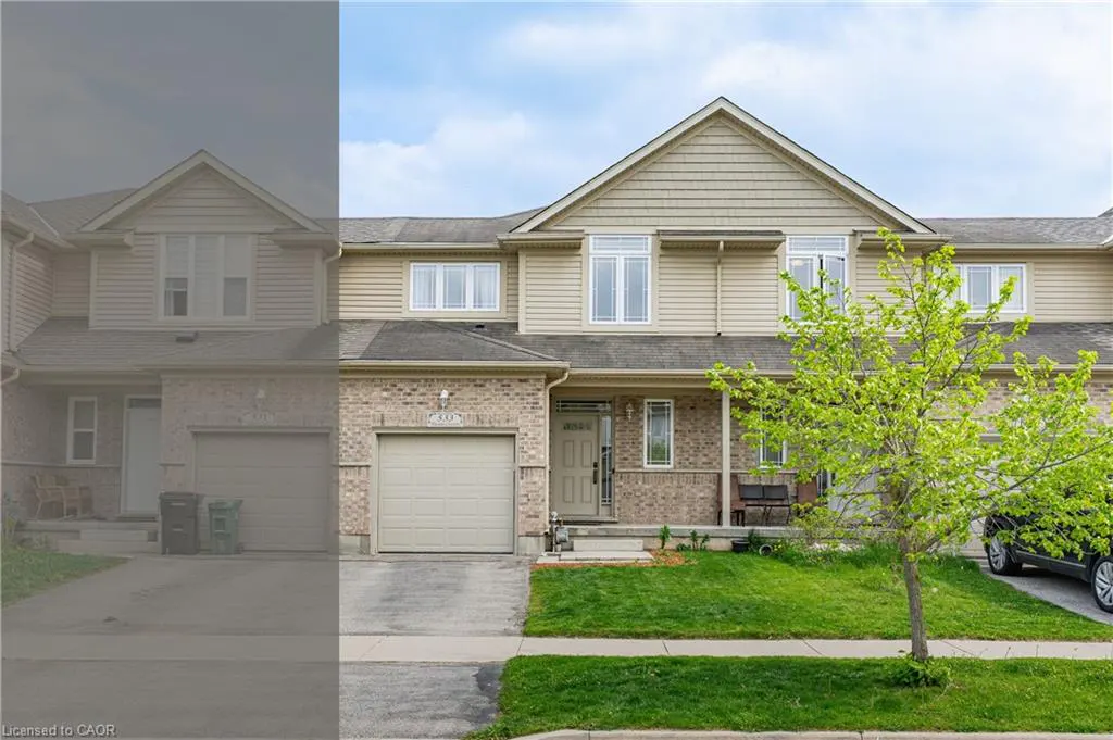 Two-story townhouse with beige siding and brick facade, featuring a garage, front lawn, and a young tree with bright green leaves.