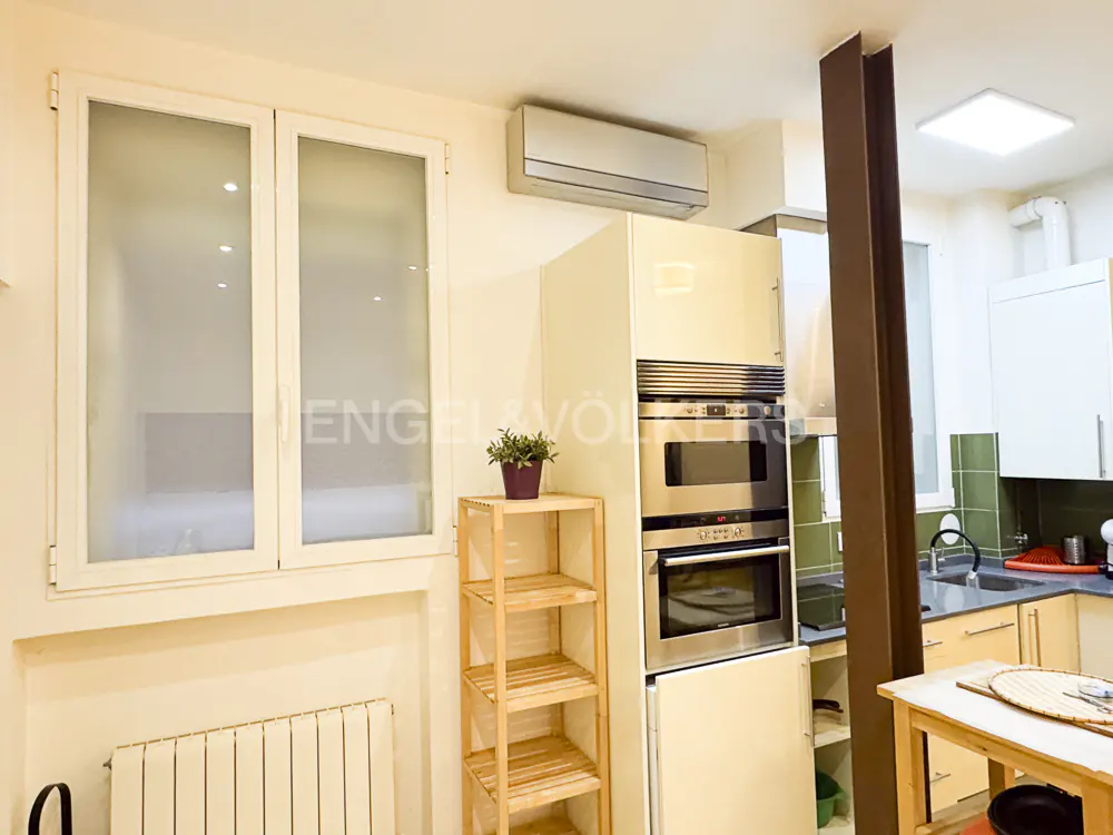 A bright kitchen with white cabinets, stainless steel appliances, and a wooden shelf with a potted plant. A large window lets in natural light.
