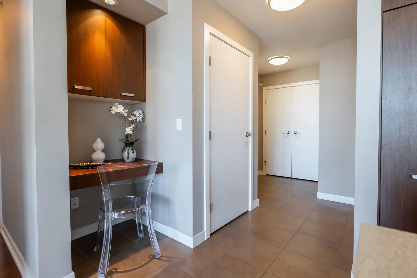 Hallway with a built-in desk and chair. The desk has a brown top and a clear acrylic chair. There is a white orchid on the desk. The floor is brown tile.