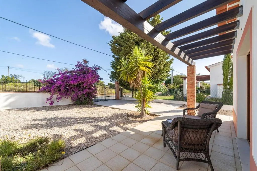 Tiled patio with wicker chairs under a wooden pergola, overlooking a gravel yard with a vibrant purple bougainvillea.