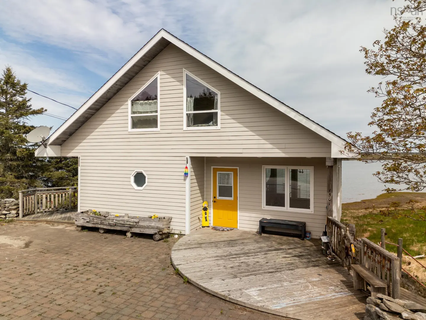 Beige house with a yellow door, triangular windows, and a round window. A wooden deck and benches surround the house. Trees and water in the background.