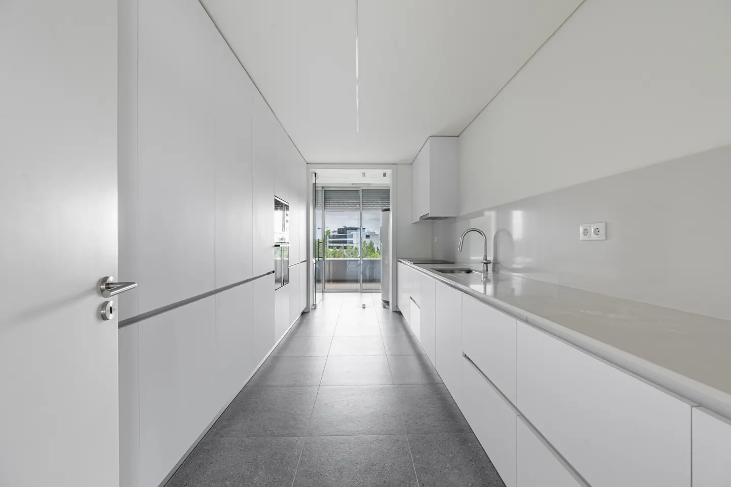Minimalist galley kitchen with white cabinets, grey tiled floors, and a glass door leading to a balcony.