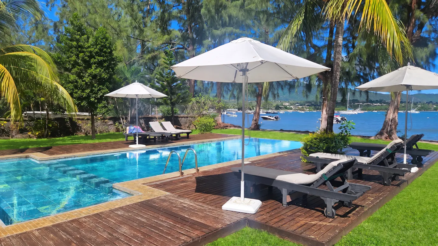 A turquoise pool with lounge chairs and white umbrellas on a wooden deck, overlooking a bay with boats and green trees.