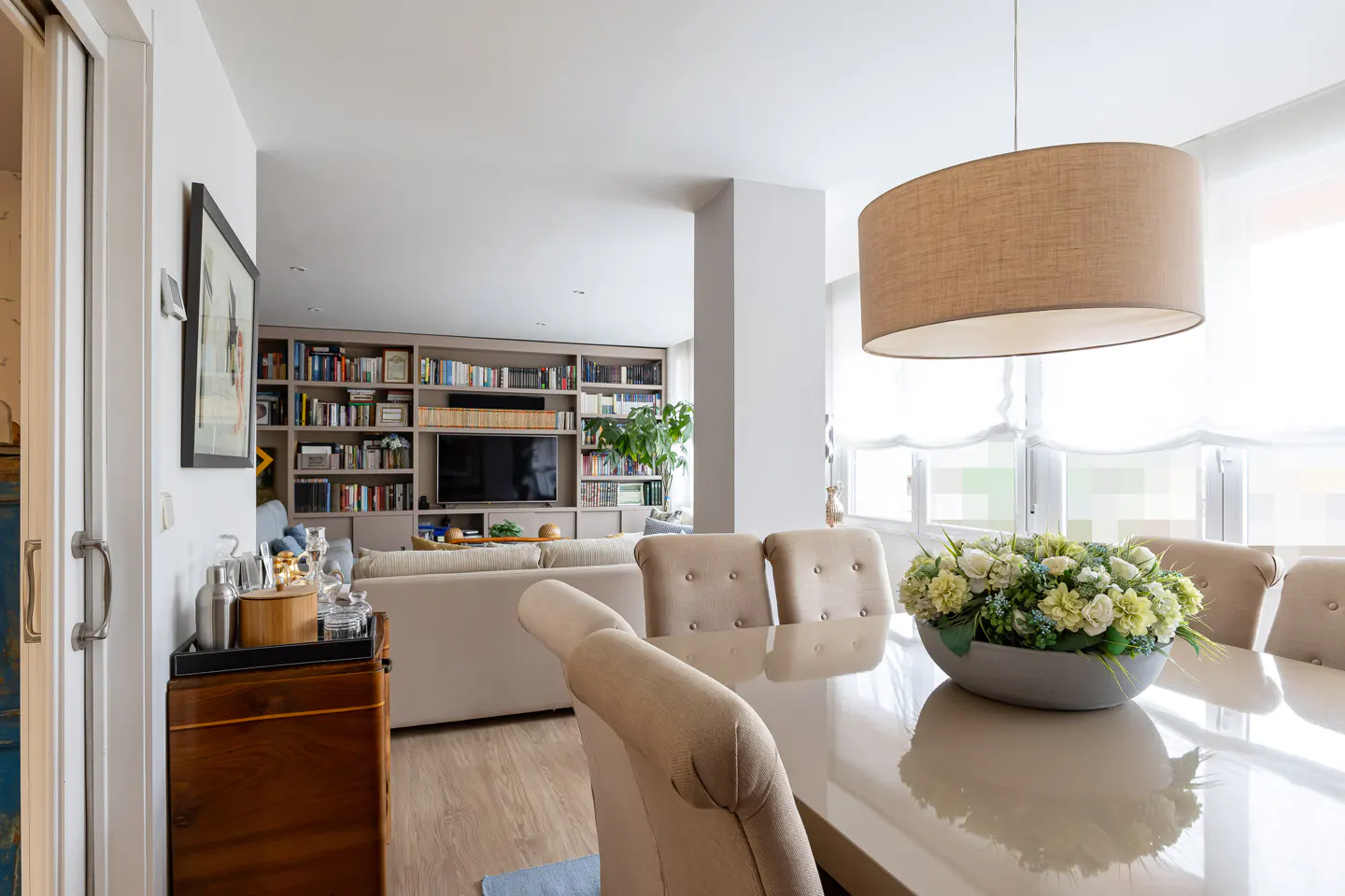 A bright, open-concept living and dining area with a large bookshelf, beige sofa, and dining table with floral centerpiece.