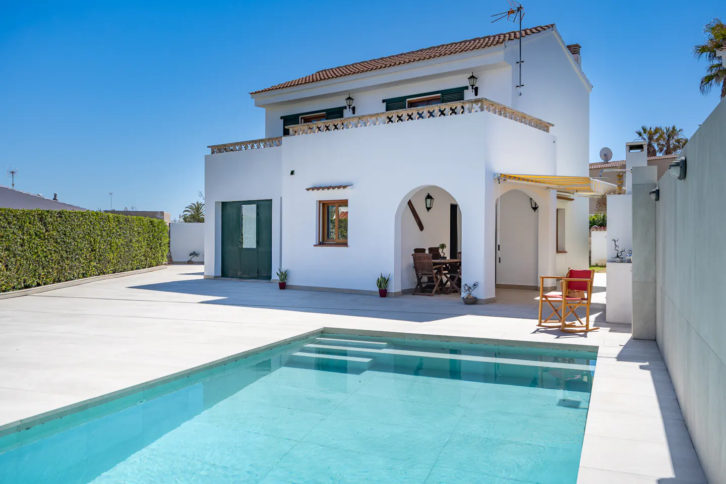 Exterior of a two-story white house with a red tile roof, a blue pool, and a green hedge under a clear blue sky.