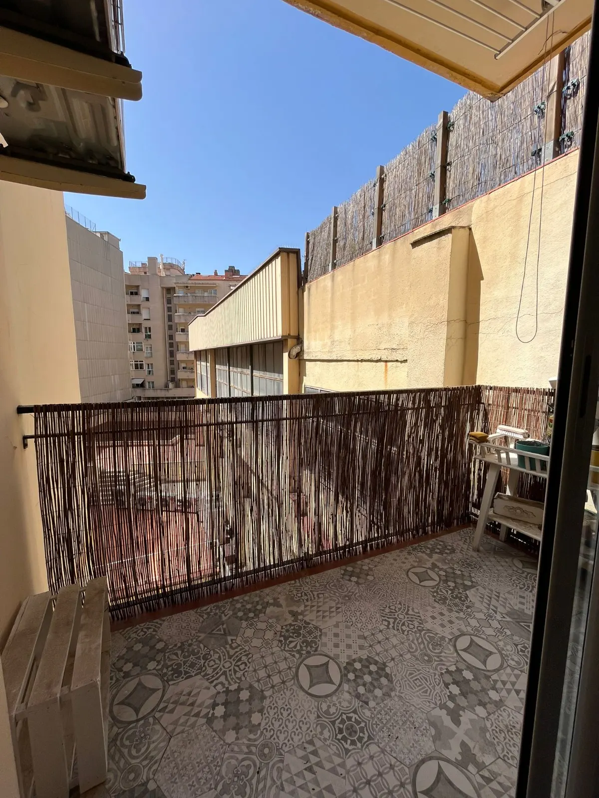 Balcony view with patterned tile floor, a bamboo fence, and a wooden bench. Buildings are visible in the background under a blue sky.