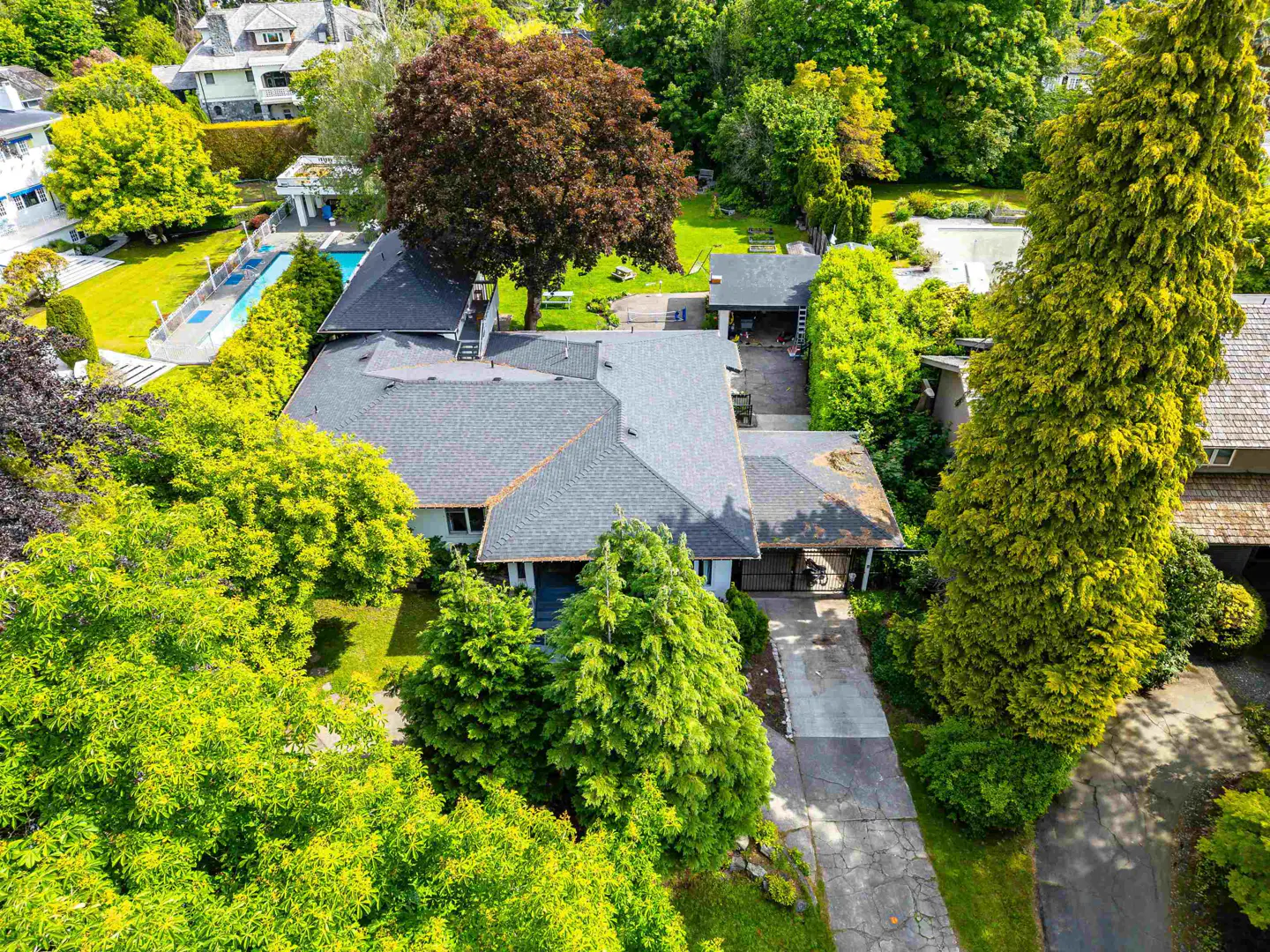 Aerial view of a single-family home with a gray roof, surrounded by lush green trees and a long driveway. A swimming pool is visible in the background.