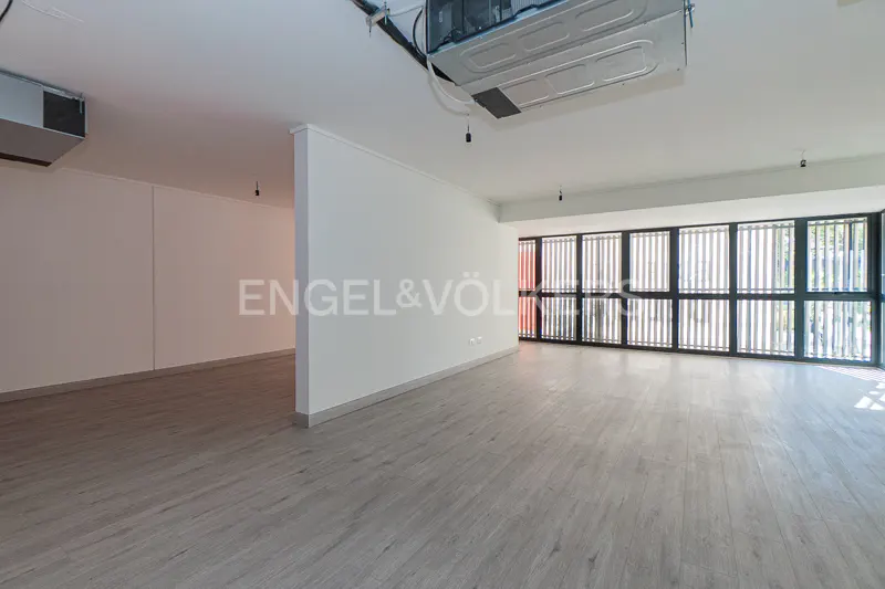 Empty room with gray wood floors, white walls, and a large window with black vertical blinds. An AC unit hangs from the ceiling.