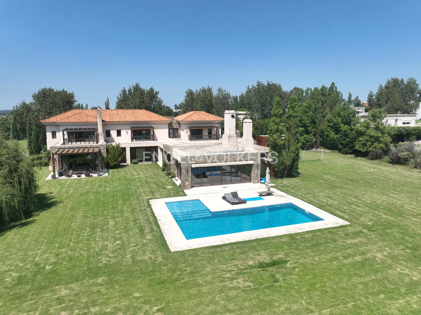 Aerial view of a large, light-colored house with a red tile roof, a pool, and a green lawn on a sunny day.