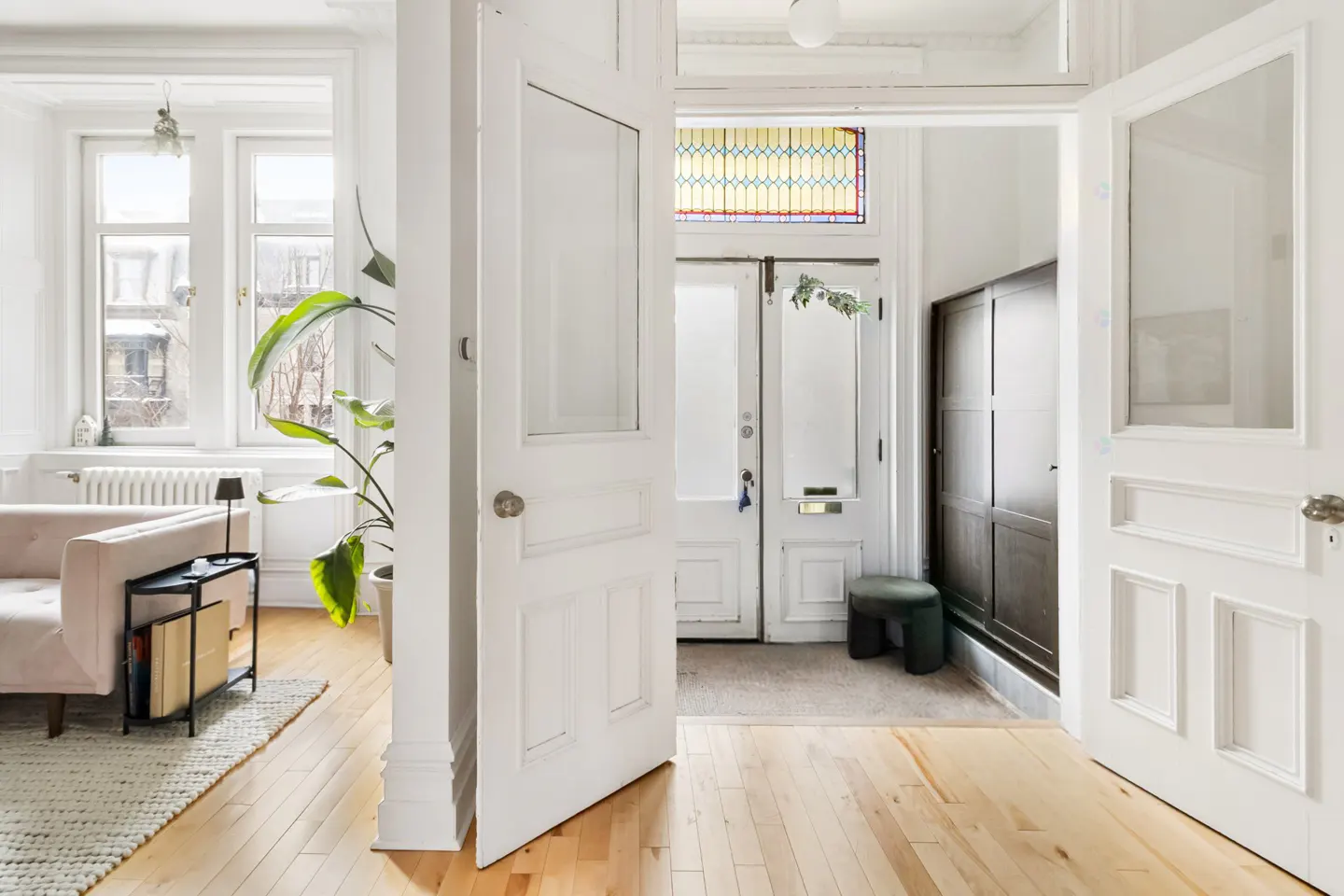 Bright entryway with open white doors, light wood floors, and a glimpse of a pink sofa and potted plant in the living room.
