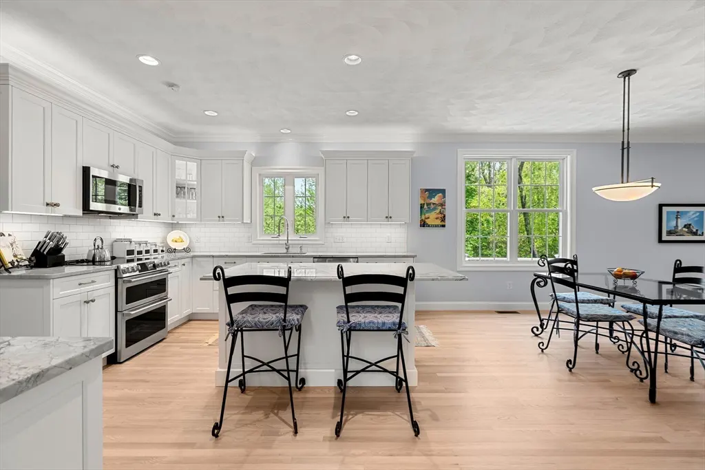 Bright kitchen with white cabinets, marble island, two black bar stools, and a dining area with a glass table.