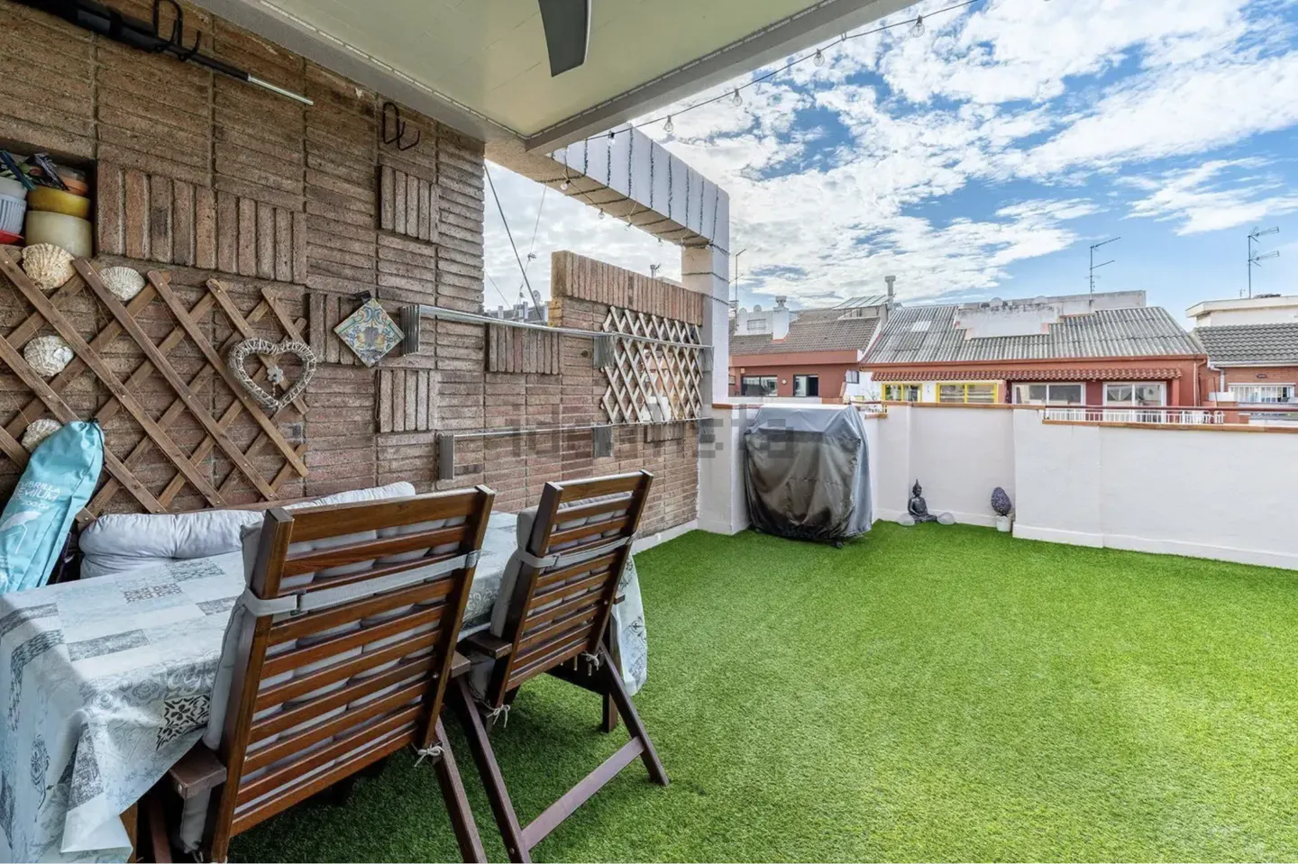 Outdoor patio with brick wall, table, chairs, and artificial grass. Grill covered in black tarp. Blue sky with clouds in background.