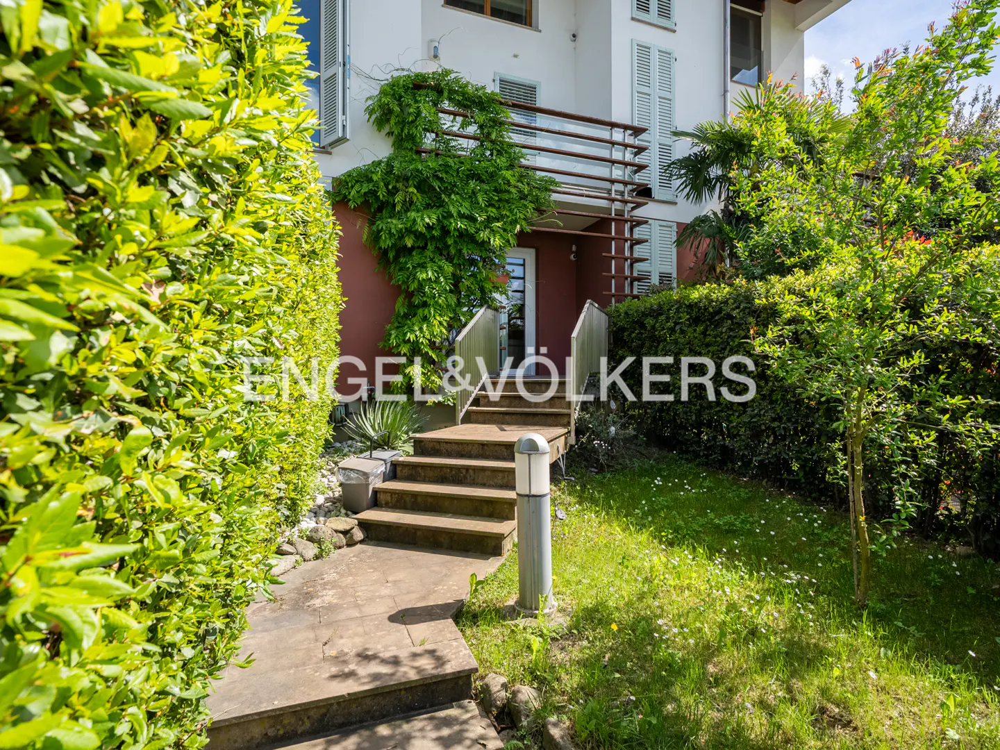 Exterior view of a two-story house with a stone walkway leading to the front door. Green bushes and trees surround the house. The house is white with red accents.