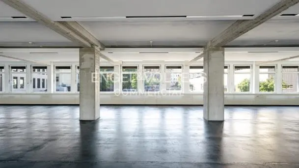 An empty commercial space with concrete pillars, beams, and a polished gray floor. Windows line the back wall, letting in natural light.