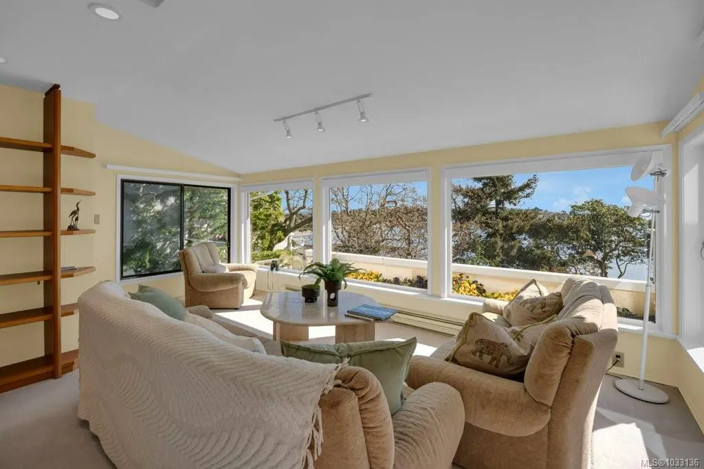 Living room with beige sofas, a tan armchair, and a light-colored oval coffee table. Large windows offer a view of trees and sky.