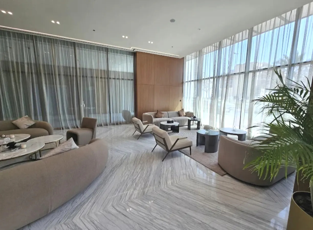 Modern lobby with beige sofas, armchairs, marble floors, wood-paneled walls, and floor-to-ceiling windows with sheer curtains.
