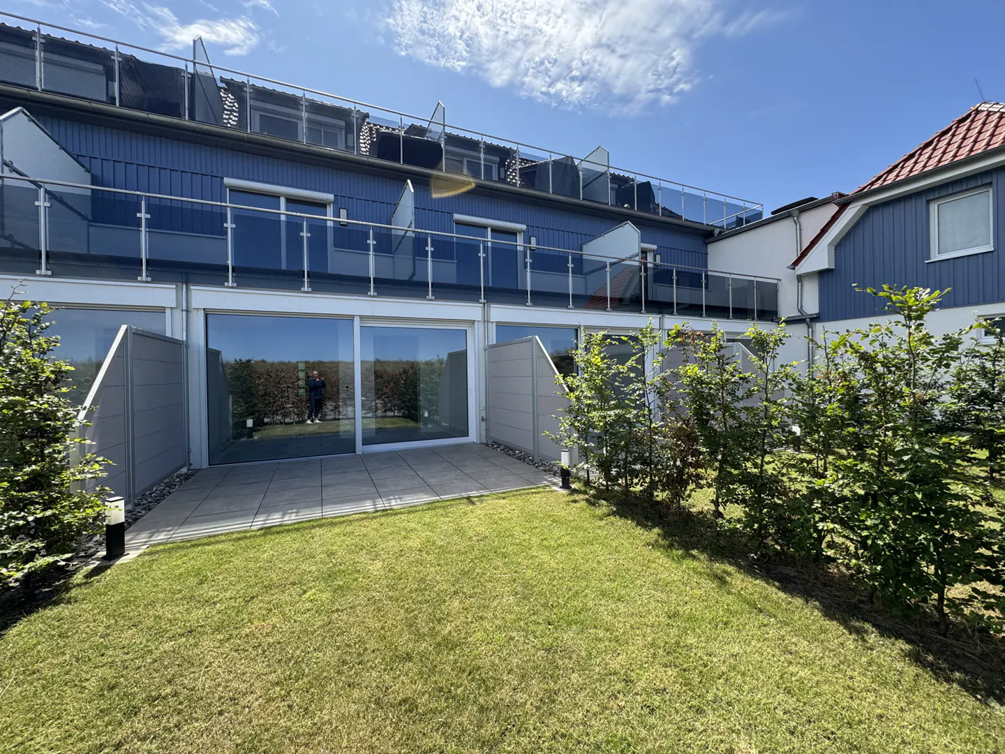 A blue building with glass balconies overlooks a green lawn and patio. Bushes line the edge of the lawn.