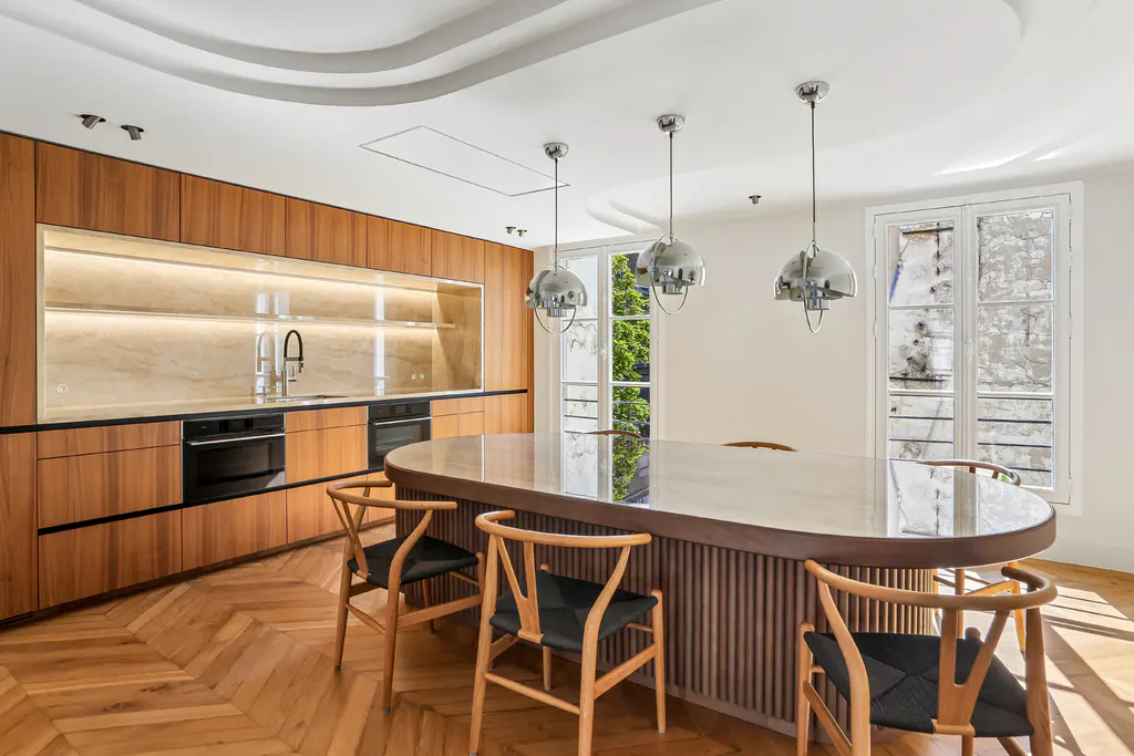 Modern kitchen with wood cabinets, a large oval island with black-seated chairs, and three chrome pendant lights.