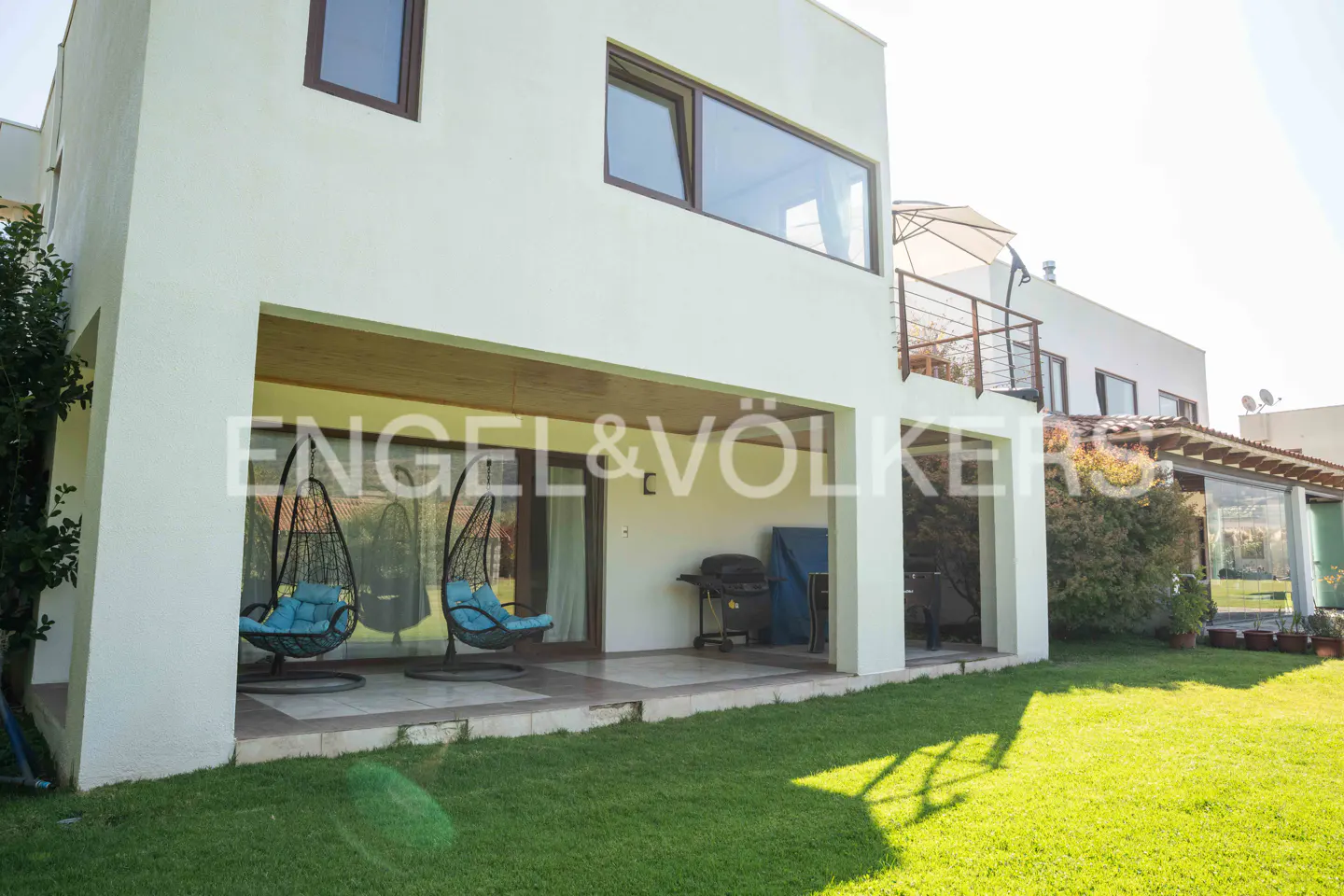 Exterior view of a modern white house with a green lawn, patio with hanging chairs, and Engel & Völkers logo.
