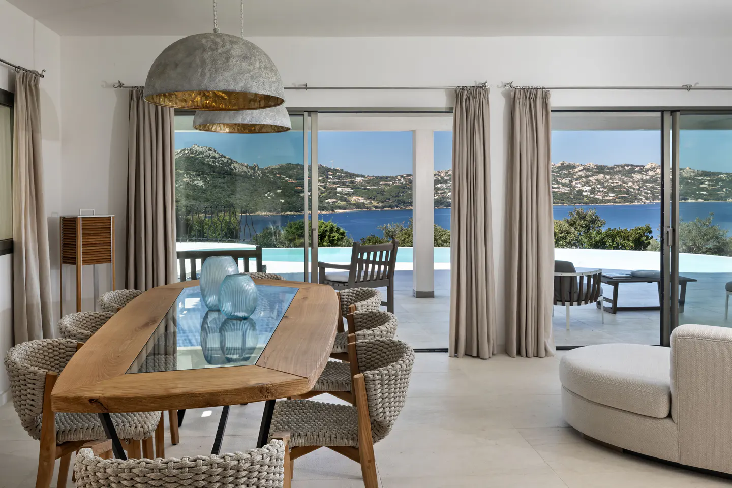 Bright dining room with a wood table, woven chairs, and blue glass vases. Sliding glass doors open to a pool and ocean view.