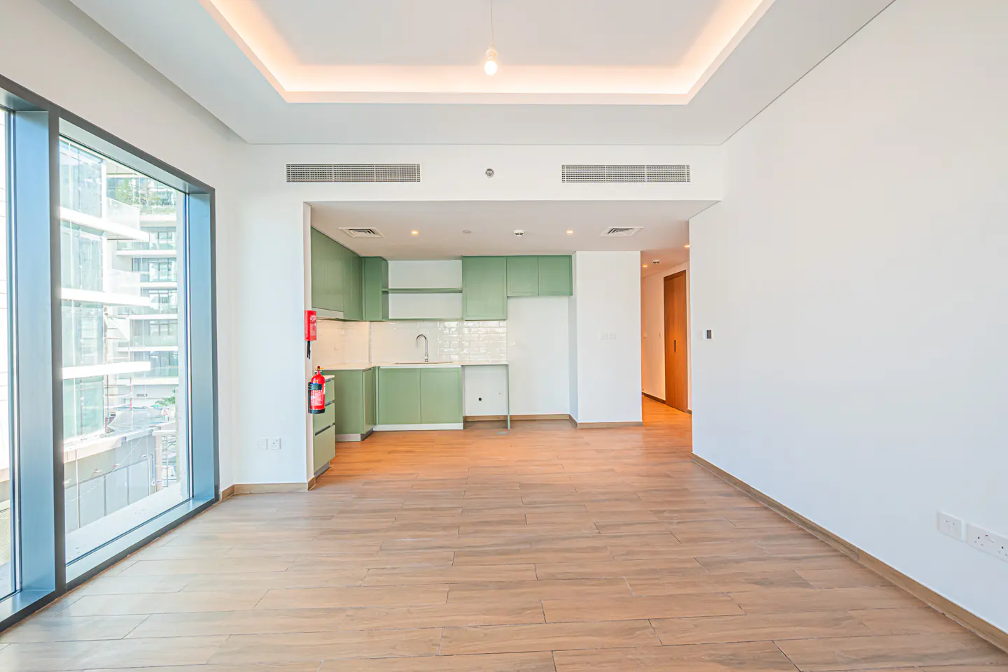 Bright, empty apartment with wood floors, white walls, and green kitchen cabinets. A large window shows a city view.