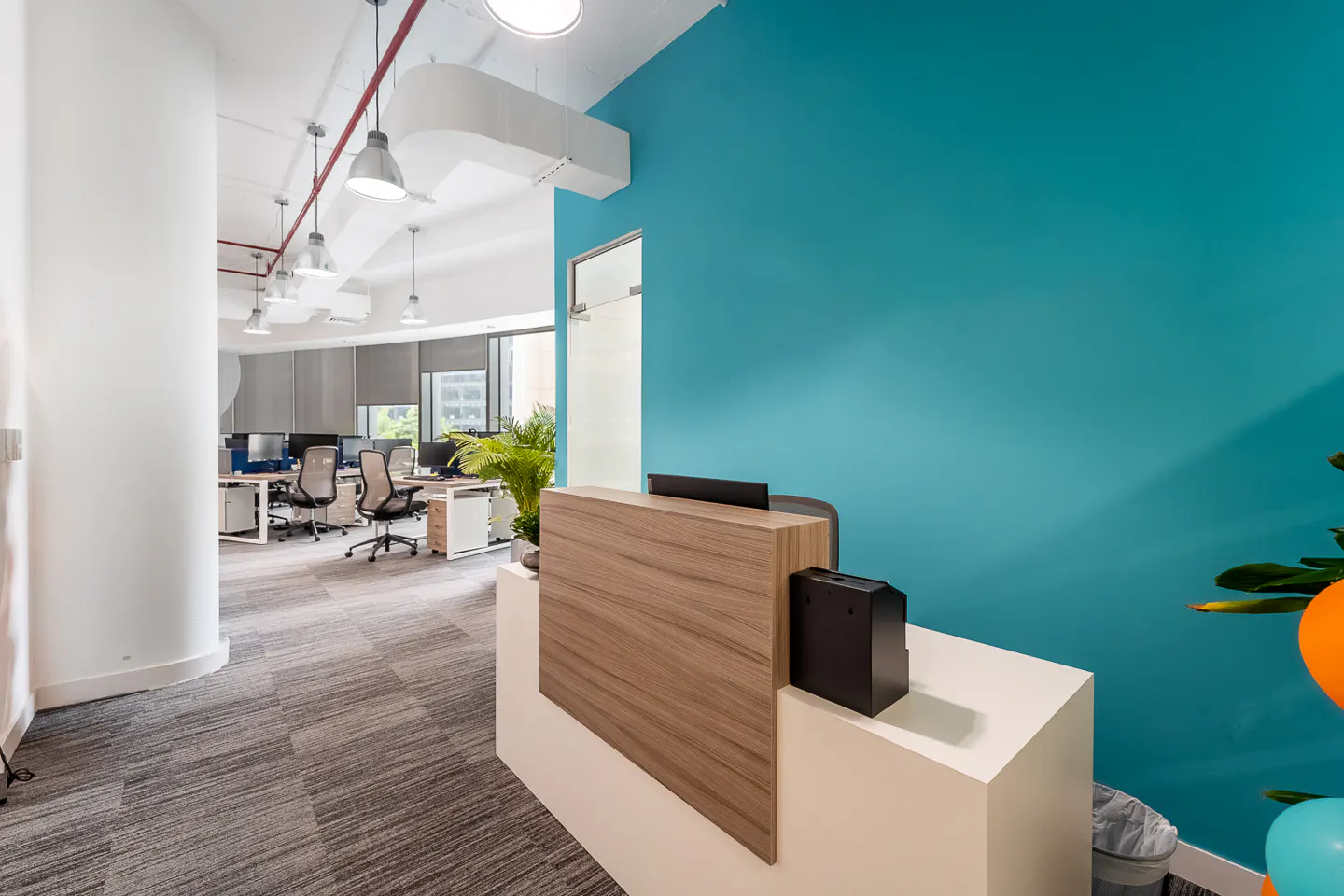Office reception area with a wood-paneled desk, white base, and a bright teal wall. Desks and chairs are visible in the background.