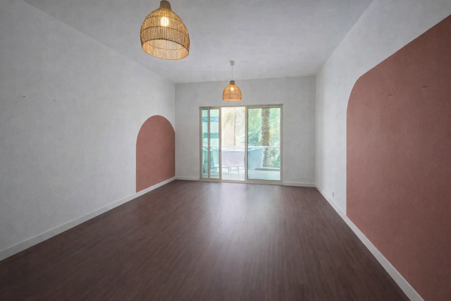 Empty room with brown wood floors, white walls with pink arches, and woven pendant lights. Sliding glass doors lead to a balcony.