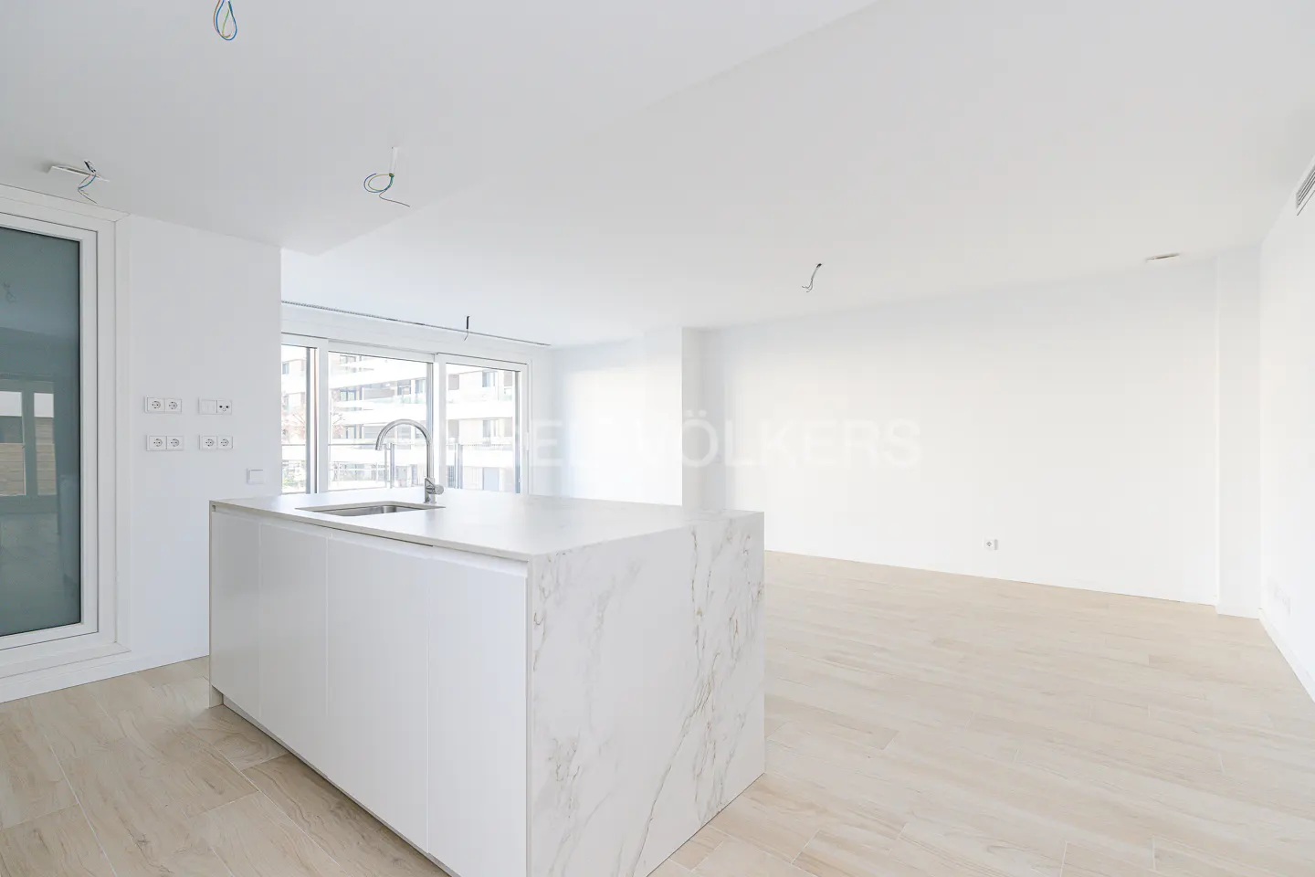 Bright, modern apartment interior with white walls, light wood floors, and a marble-topped kitchen island with a sink. Large windows provide natural light.