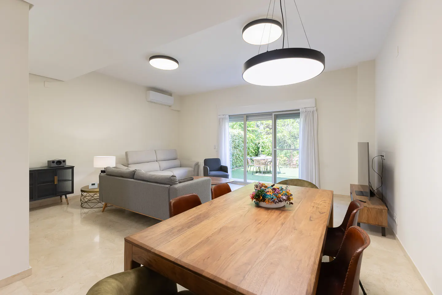 Modern living room with a wooden dining table, leather chairs, grey sofa, and sliding glass doors leading to a patio.