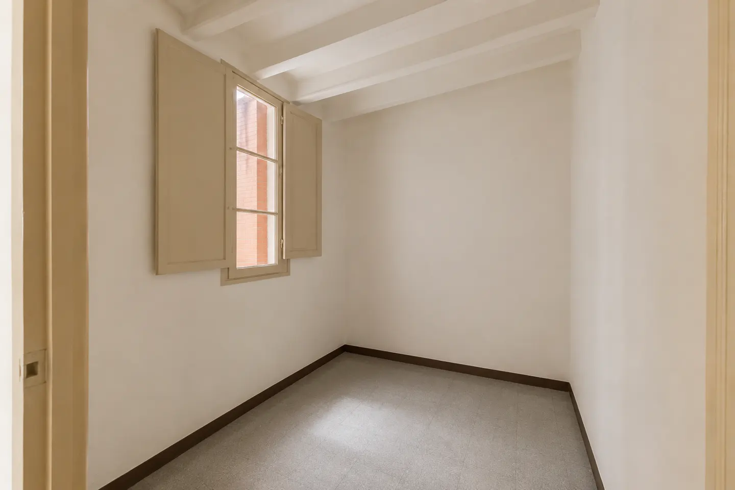 Empty room with white walls, gray carpet, and a window with beige shutters. White ceiling beams are visible.