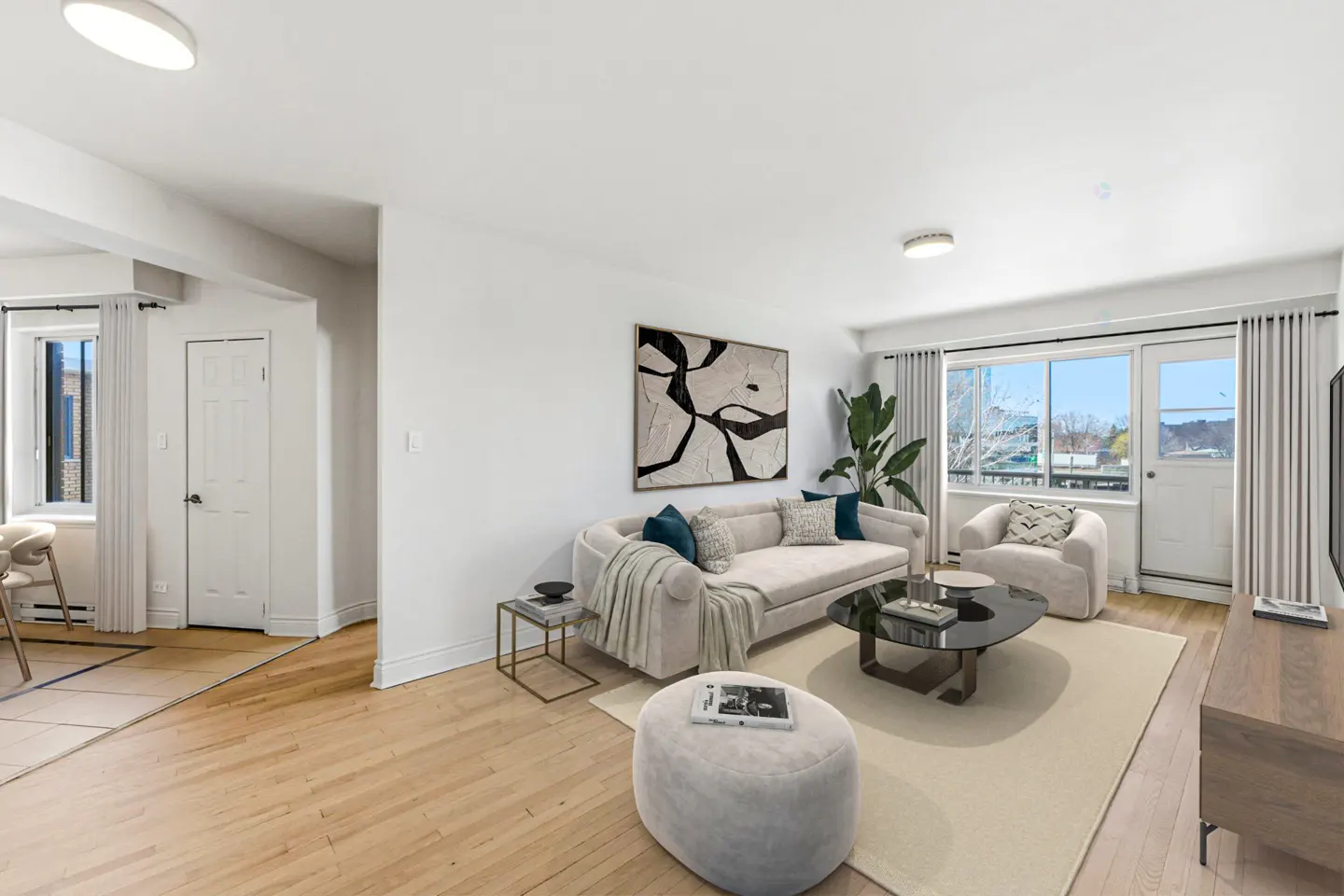 Bright living room with hardwood floors, white walls, and neutral furniture. Abstract art hangs above a sofa with blue pillows. Balcony access visible.