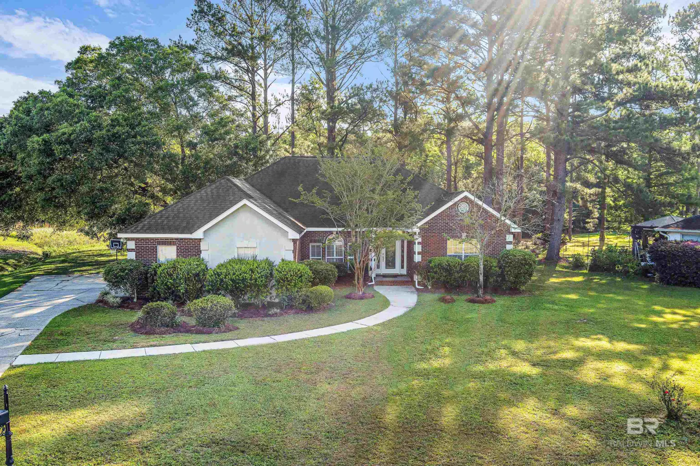 A single-story brick house with a gray roof, white trim, and a curved walkway leading to the front door. The house is surrounded by green grass and trees.
