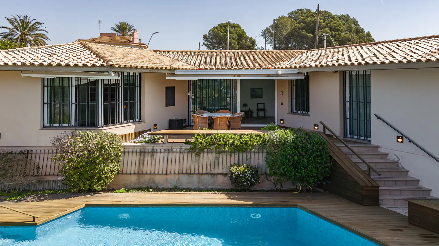 Backyard view of a beige house with a terracotta tile roof, a blue pool, and a wooden deck.