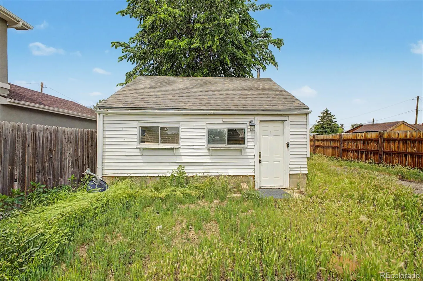 Small white detached garage with a shingled roof, two windows, and a white door, set in a yard with tall, overgrown grass.