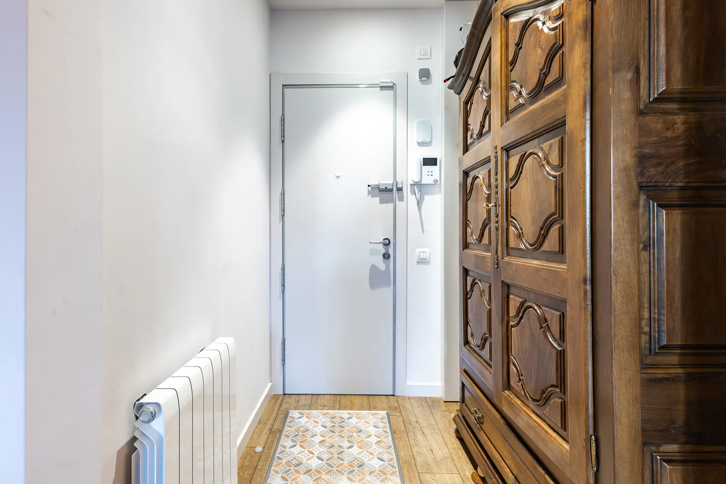 Entryway with white walls, a white door, and a large, dark wood armoire. A radiator sits near the wall, and a patterned rug lies on the wood floor.
