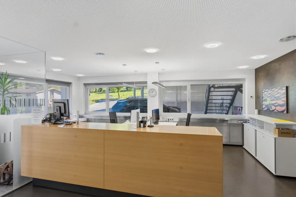 Modern office reception with a large light wood desk, white cabinets, and large windows overlooking a green outdoor area.