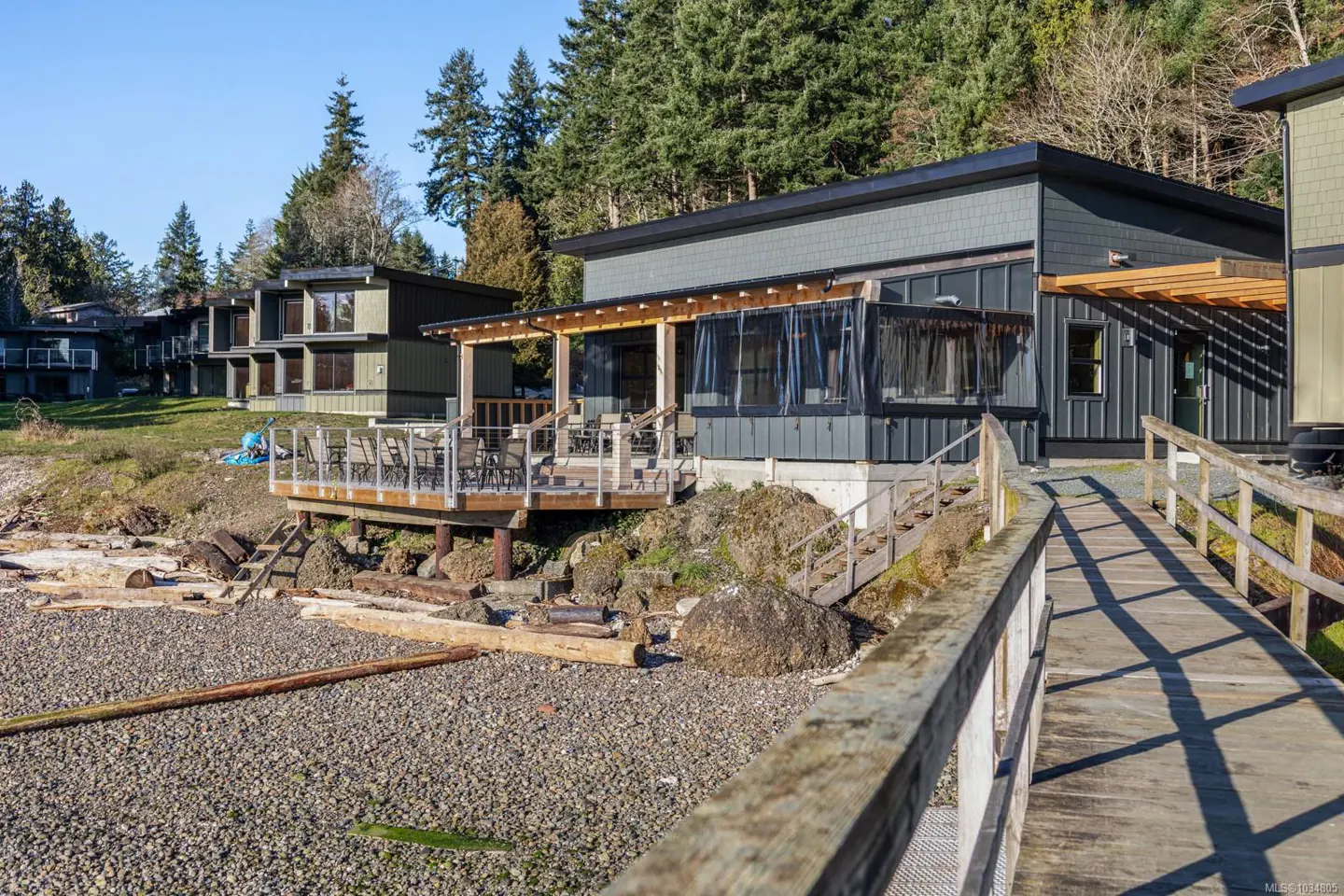 Exterior view of modern gray cabins with wooden decks on a rocky beach, connected by a wooden walkway. Trees in background.