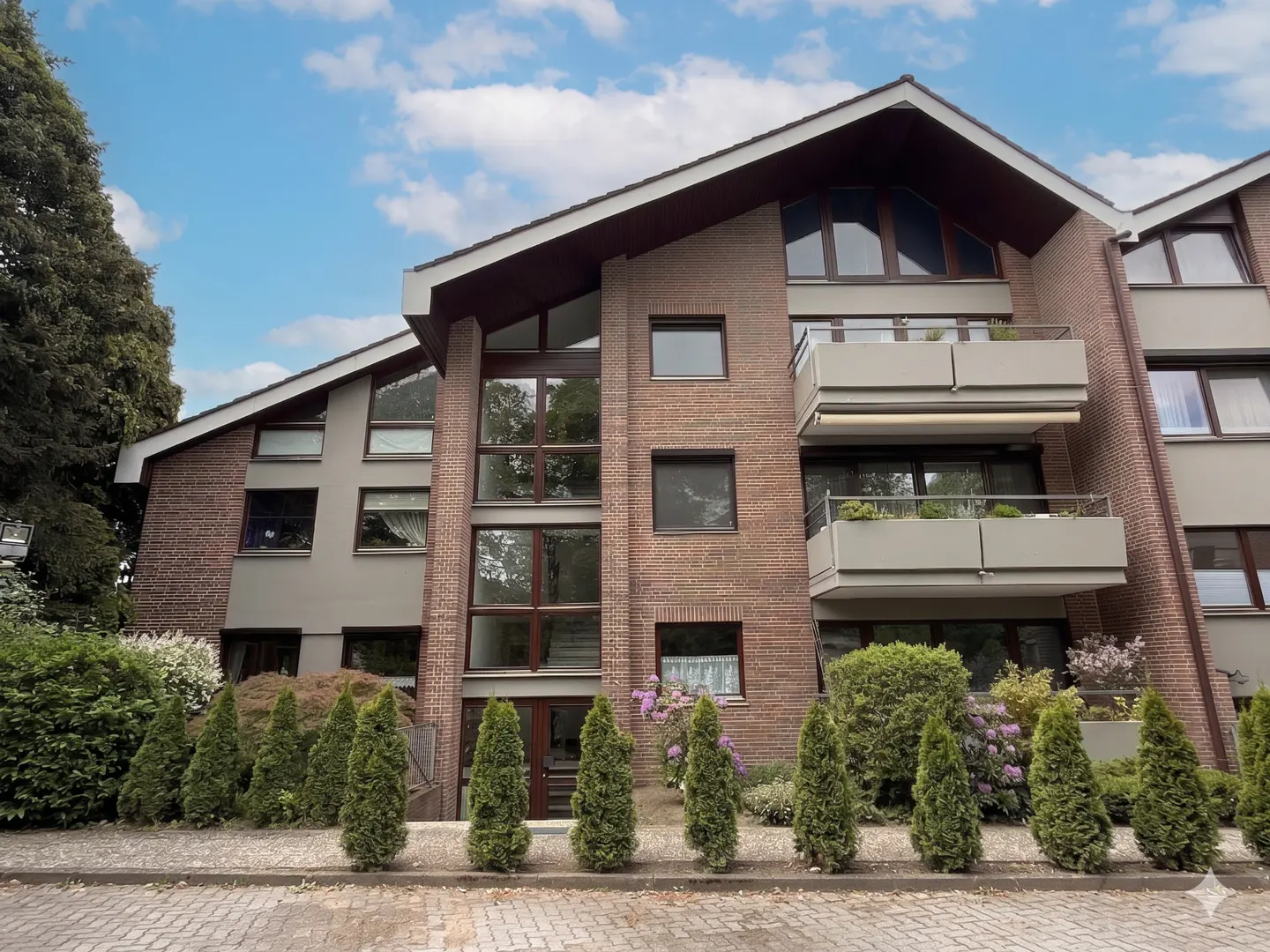 Exterior view of a brick apartment building with balconies, green shrubs, and a blue sky.