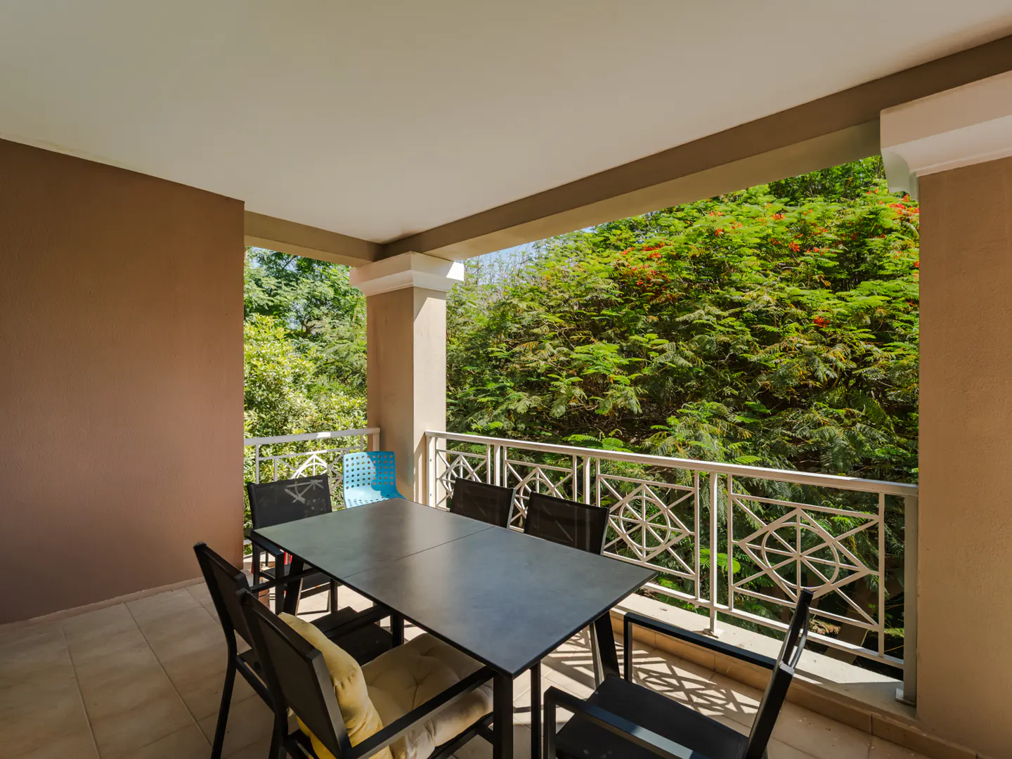 Covered patio with a black table and chairs. White railing overlooks lush green trees. One blue chair is visible.