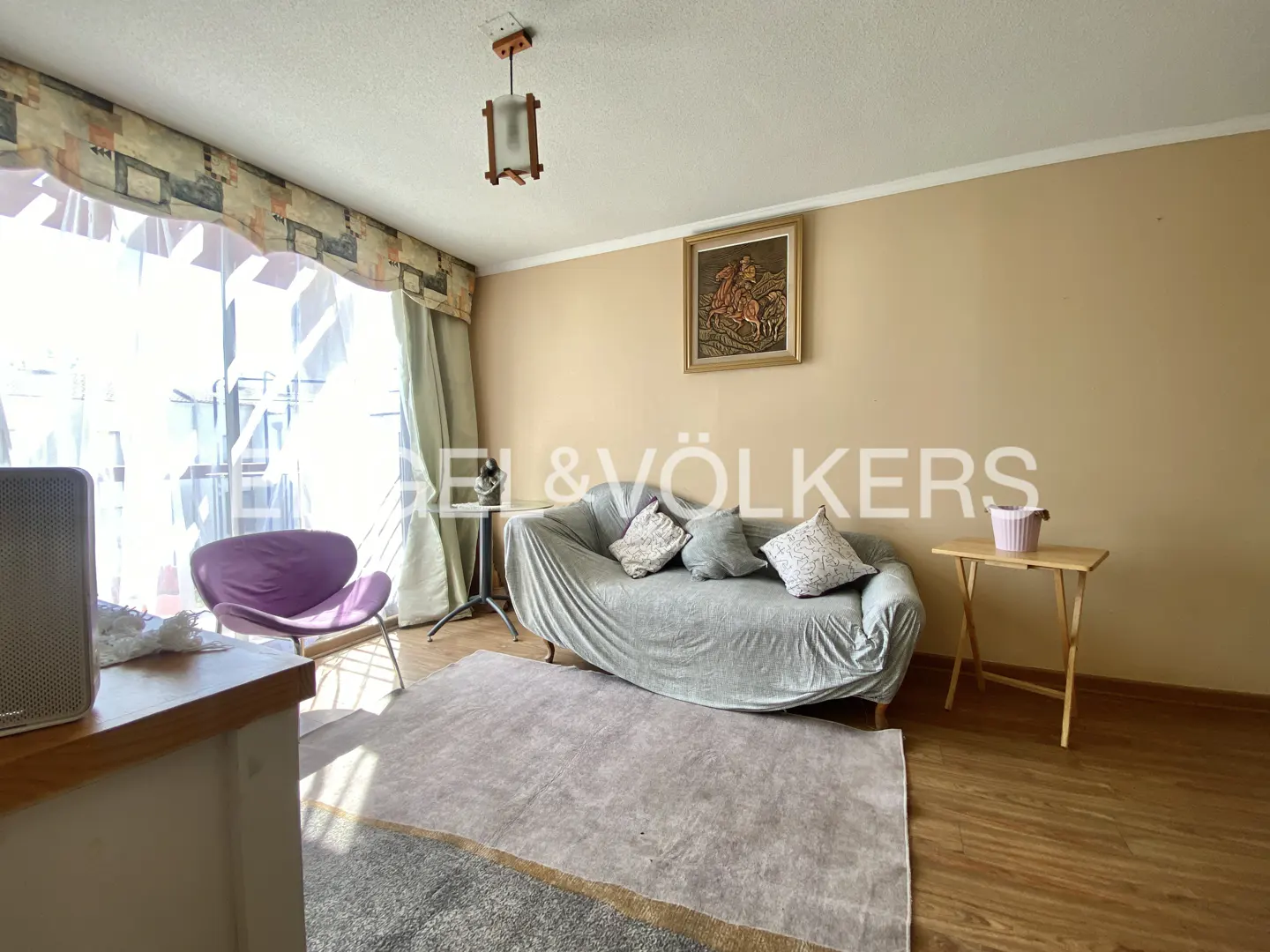 Living room with beige walls, wood floors, a gray sofa with pillows, and a purple chair near a bright window.