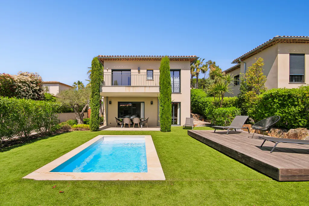 Two-story beige house with a pool, lawn, and wooden deck with lounge chairs under a clear blue sky.