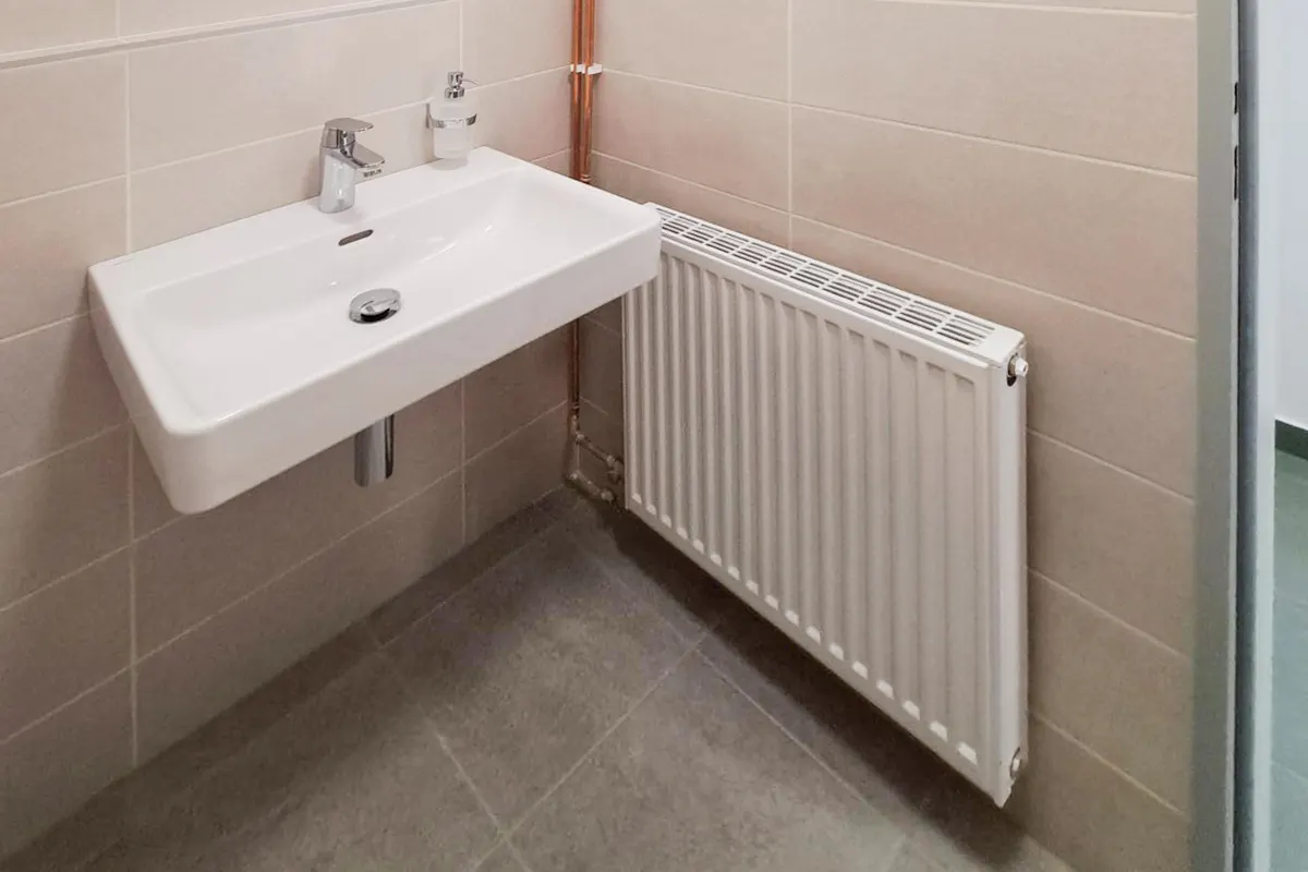 Bathroom corner with a white sink, chrome faucet, and soap dispenser. A white radiator is on the wall next to the sink. Beige tiled walls and gray tiled floor.