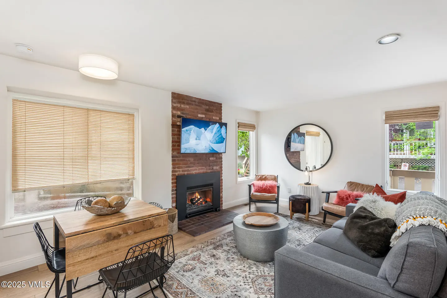 Cozy living room with gray sofa, brick fireplace with TV, round mirror, and wooden dining table near a window.