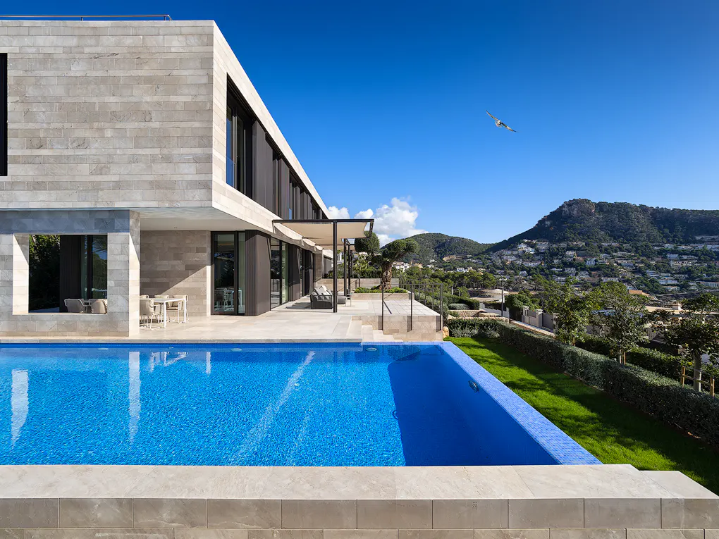 Modern stone house with a blue pool, patio, and mountain view under a clear blue sky.