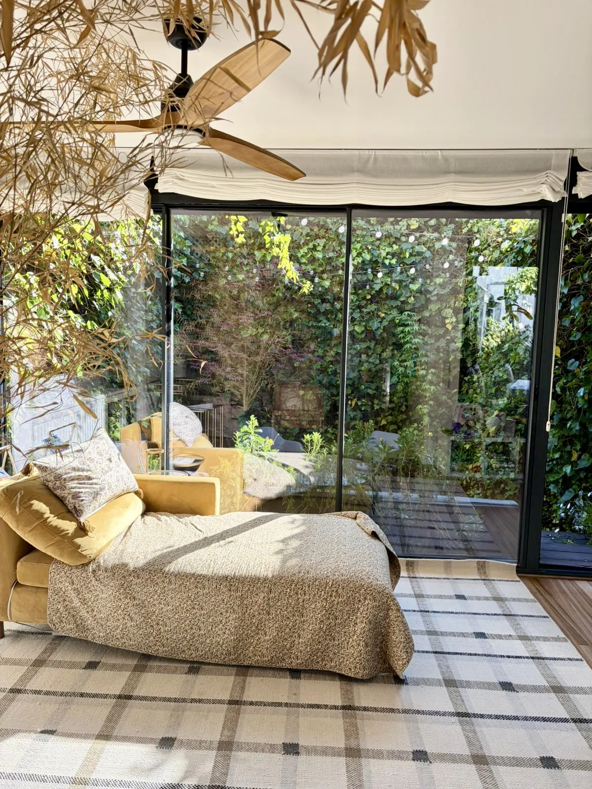 Sunroom with a yellow chaise lounge, plaid rug, and large windows overlooking a lush green garden. A ceiling fan with wooden blades is visible.
