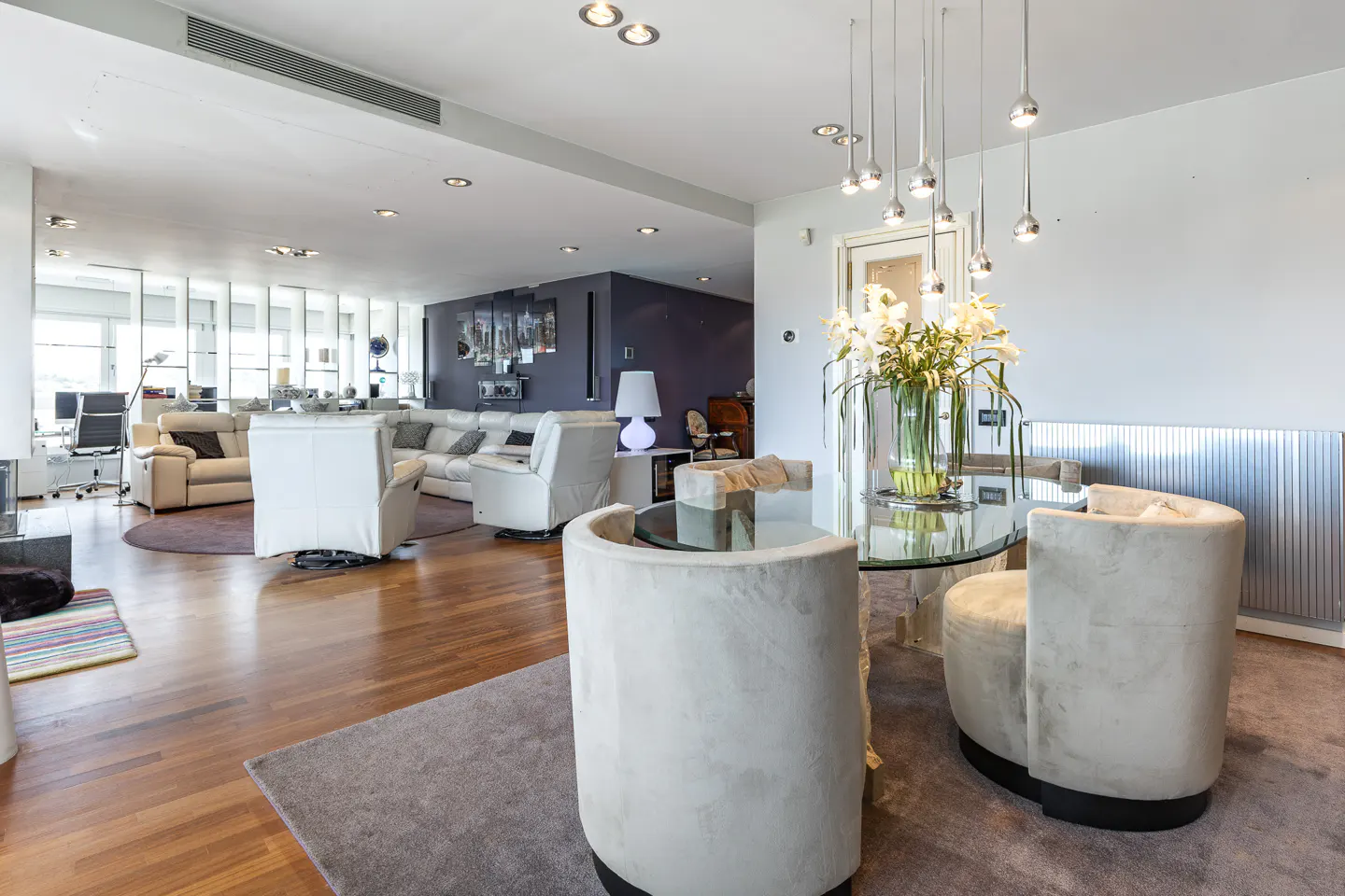 Open-concept living and dining area with hardwood floors, white sofas, and a glass dining table with white chairs.